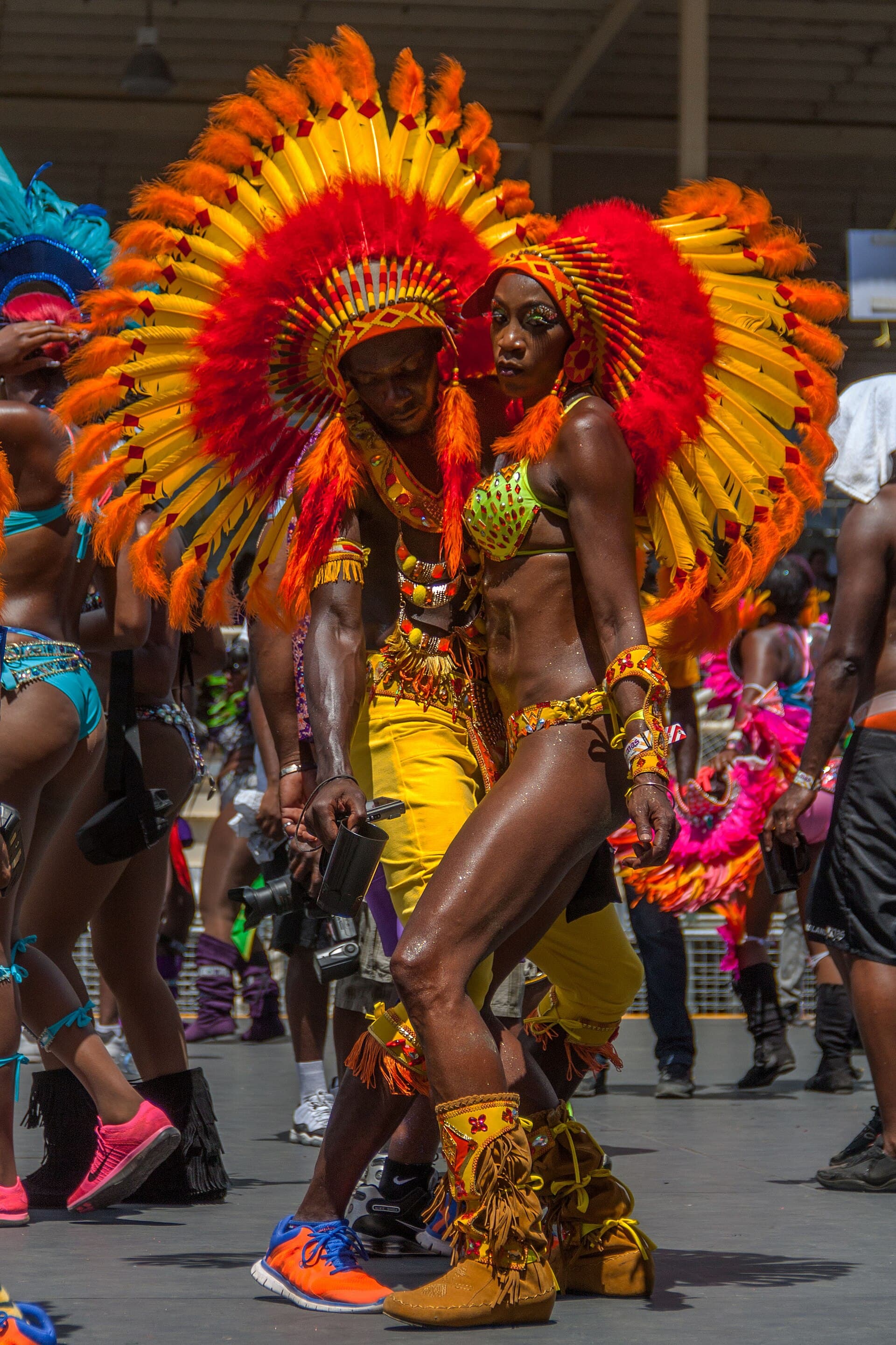 Two masqueraders in huge fanned orange, yellow and red feathered headdresses and beaded costumes dancing during the Carnival Tuesday Parade of the Bands, with other revellers behind them