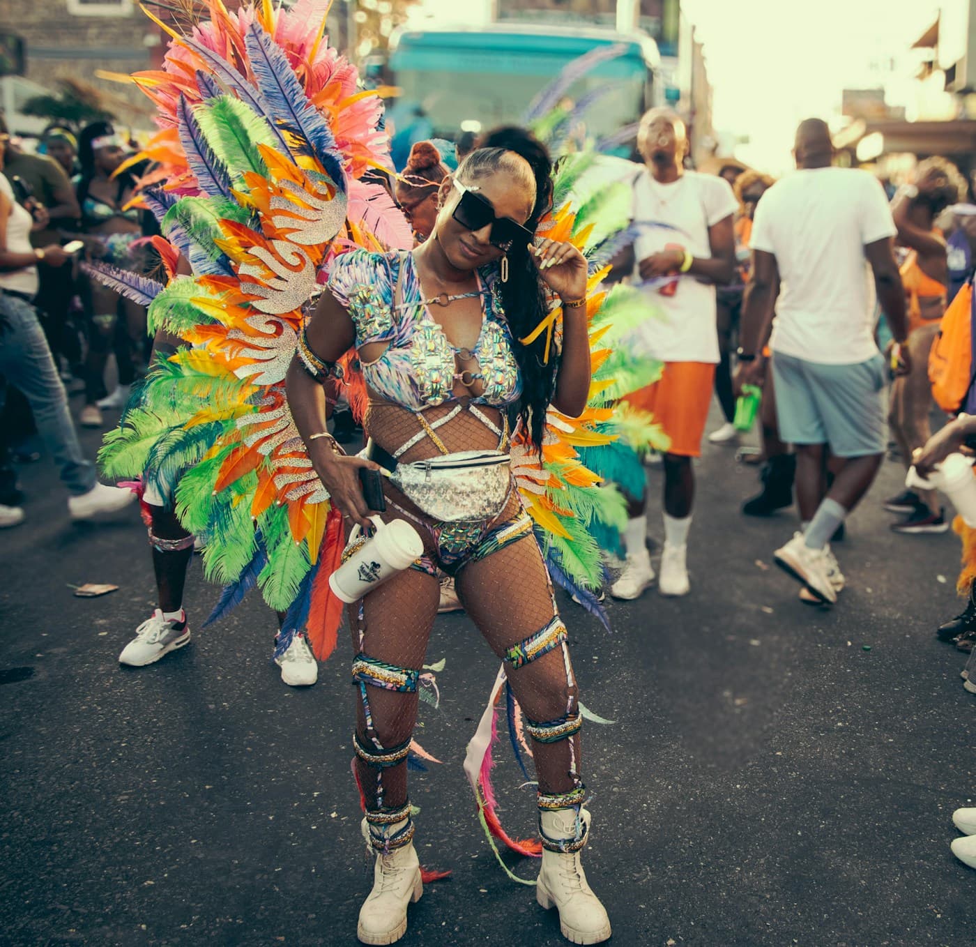 A woman in sunglasses wearing a beaded costume with a large rainbow-coloured feather backpack poses on a busy street during Carnival
