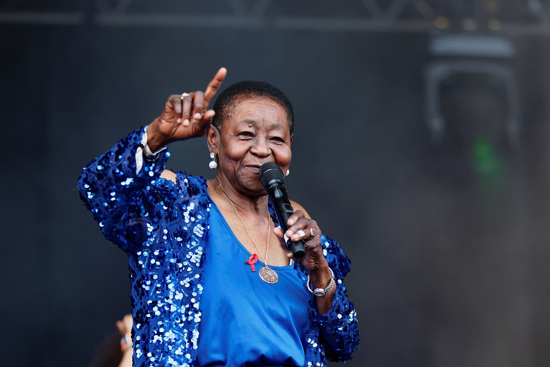 Calypso Rose in a sparkling blue jacket, pointing skyward as she sings at the Vieilles Charrues festival in France in 2016