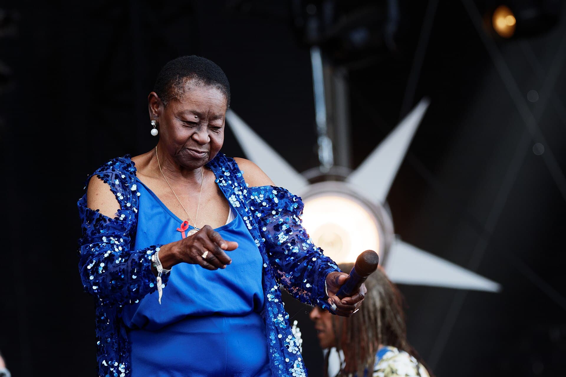 Calypso Rose performing on an outdoor festival stage in 2016, wearing a blue top and a blue sequinned jacket, microphone in hand and smiling as she dances.