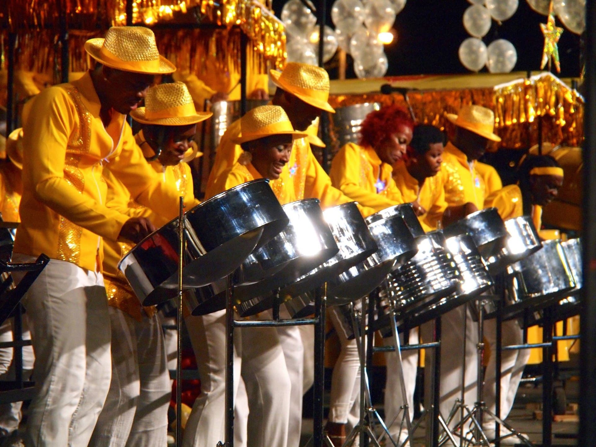 A steel orchestra in matching yellow shirts and straw hats plays chrome steelpans on a lit stage at night during Trinidad and Tobago Carnival.