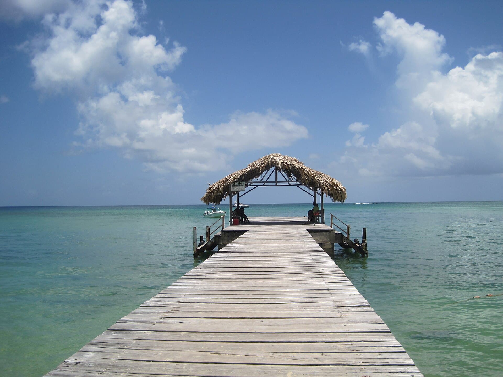 Long wooden jetty with a thatched-roof hut at the end stretching into calm turquoise water at Pigeon Point, Tobago, under a blue sky with white clouds