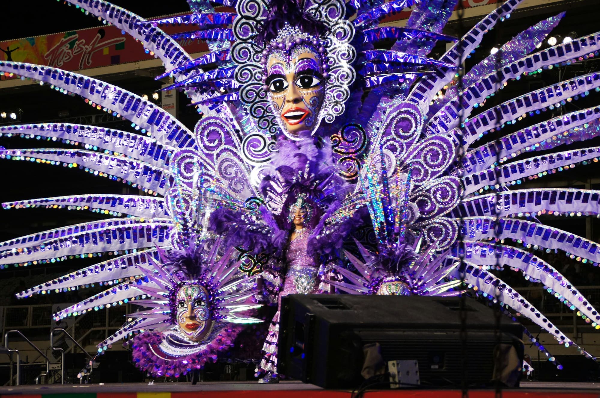 A huge king or queen style Carnival costume in purple, lilac and silver with fanned spikes, swirls and painted masks spreads across a stage at night in Port of Spain, the wearer at its centre (photo from a past Carnival)