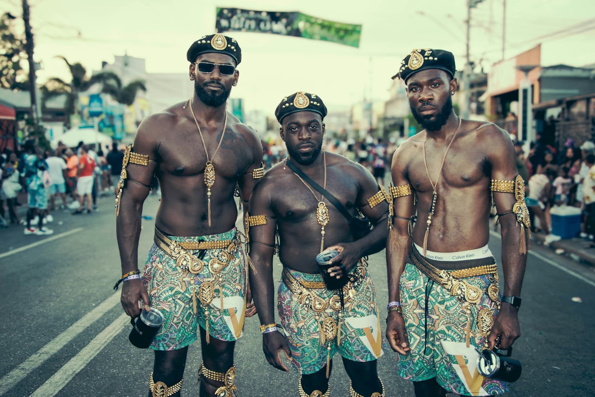 Three bare-chested men in matching patterned shorts, peaked caps and gold arm cuffs stand side by side on a street lined with crowds at Trinidad and Tobago Carnival 2024